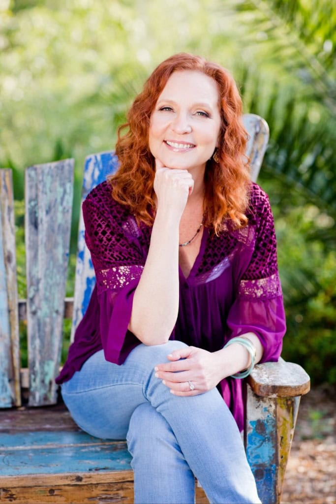 Kari Samuels, a smiling intuitive counselor with red hair, sitting on a rustic bench outdoors surrou.